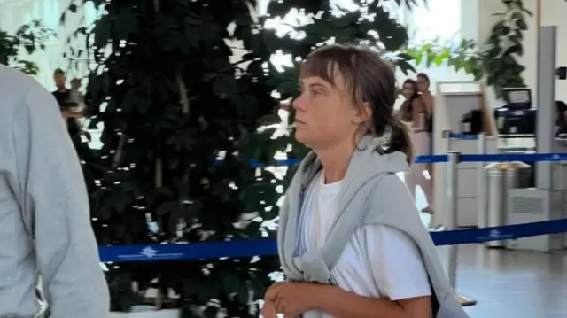 Greta Thunberg wearing a grey tracksuit walking through what appears to be an airport