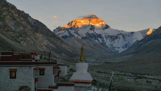 Tibetan structures in the foreground, in the background is a snow-capped mountain lit by the sun