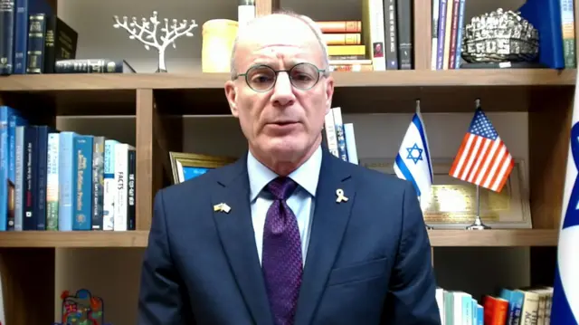 Yachiel Leiter in dark suit and purple patterned tie sits down in from of a wooden bookcase