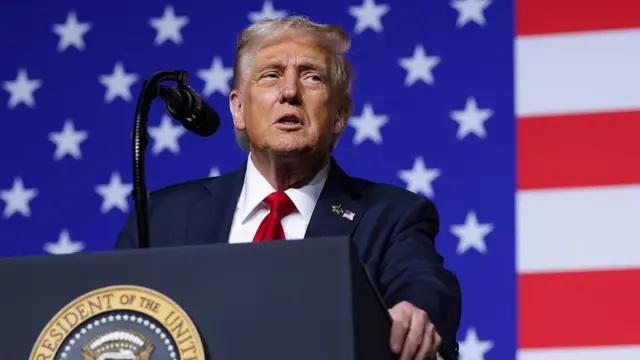 Trump speaking into a conference mic in front of a large US flag