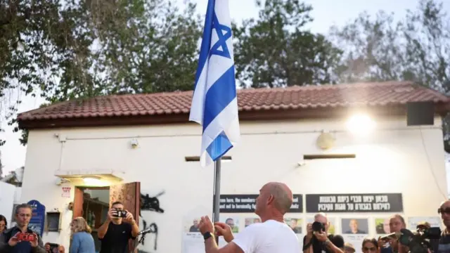A member of the kibbutz community of Kfar Aza lowers an Israeli flag to half mat 7 Oct 2025