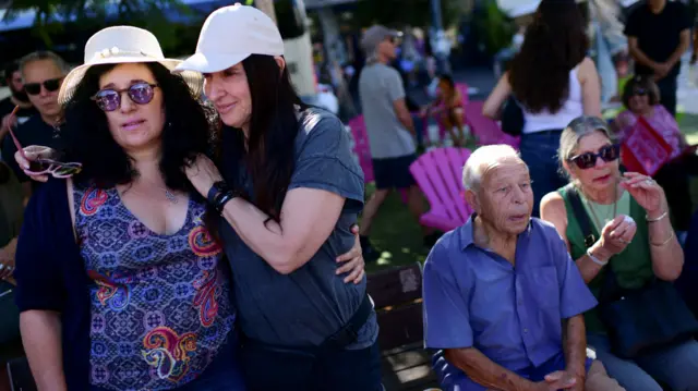 Two women hugging and people sitting on chairs next to them and behind them at a memorial outdoors