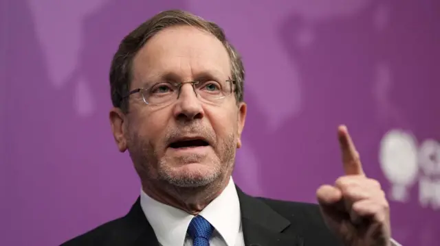 Herzog with his left index finger raised as he talks. The background is purple