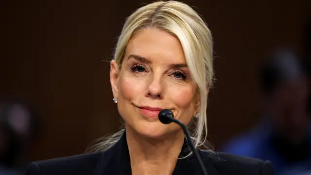 Pam Bondi in front of a microphone in Congress. She is smiling and is wearing a black jacket.