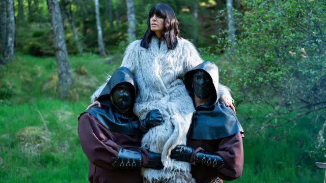 Claudia Winkleman in faux fur coat on the shoulders of two masked men in a field as she introduces a challenge on The Traitors