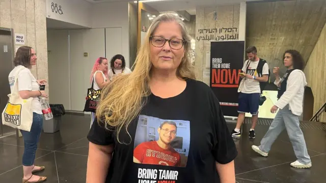 A woman wearing a T-shirt that displays a picture of a younger man with the words "bring Tamir home". She is looking at the camera, and people are walking past her in the background