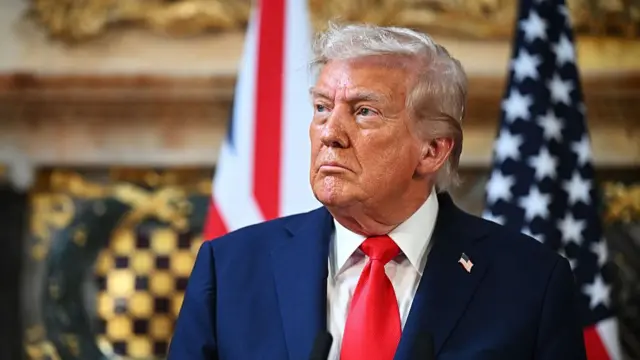 Trump in a suit and tie looking towards his right, stood in front of two US confederate flags