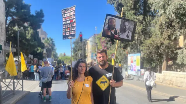 Bar and Atalia Regev stand facing the camera holding up signs showing images of the hostages