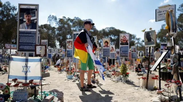 A man draped in a German flag and holding an Israeli flag stands among signs that have been put up to commemorate the victims of the attack