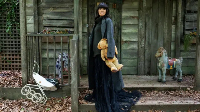 Claudia Winkleman stands outside a derelict shed in a traitors mission holding an old teddy bear