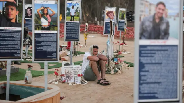 A man sits in a large display, with a white fabric on his head. His arms are folded around his knees, as he looks on solemnly.