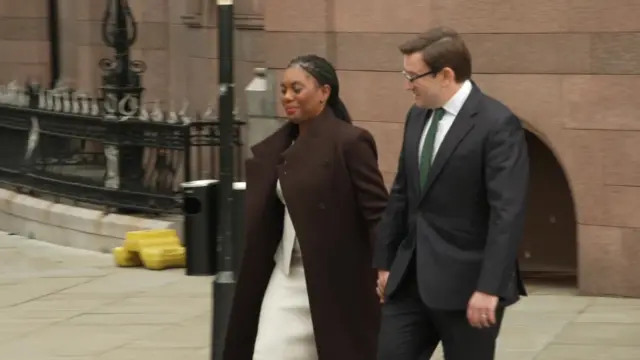 Kemi Badenoch pictured wearing a brown coat. She is with her husband, Hamish, who is wearing a black suit and green tie.