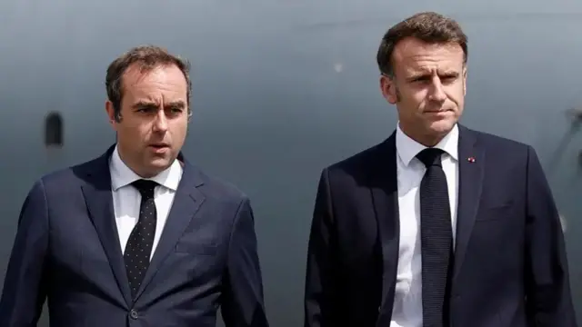 Sébastien Lecornu, who has dark hair and is wearing a dark suit, looks off to the centre while standing next to French President Emmanuel Macron, who is also wearing a dark suit.