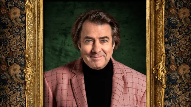 Jonathan Ross in the show's official portrait, smiling and wearing a black top and salmon coloured blazer