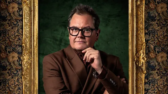 Alan Carr standing inside a golden photo frame with his left hand to his chin