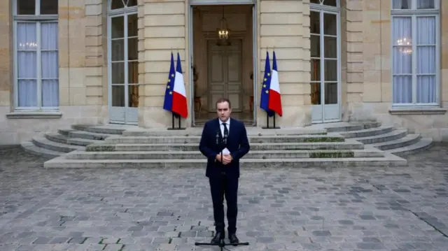 Sébastien Lecornu stands in a courtyard, in front of a microphone, with the French and EU flags behind him