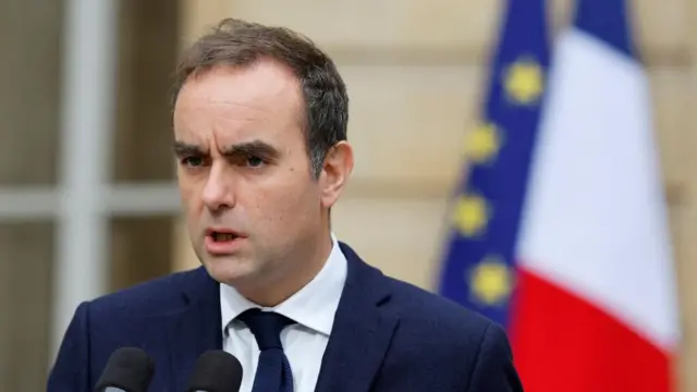 Lecornu speaking into a microphone earlier today, he is wearing a suit and the French and EU flags can be seen behind him
