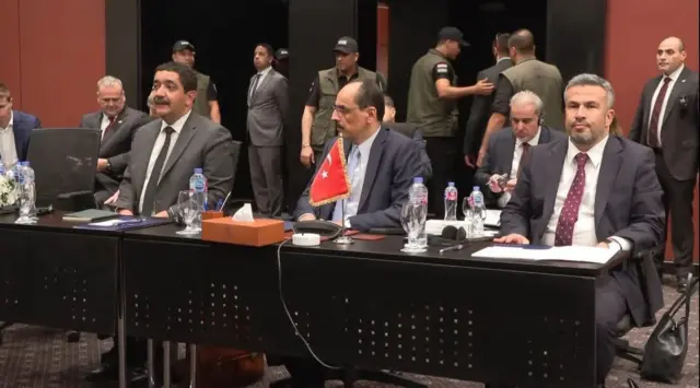 Three men in suits sit a table that has a small Turkish flag on it.