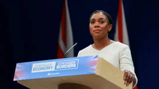 Badenoch speaking at party conference in front of Union Jack flag, at a lectern which ready "STRONGER ECONOMY STRONGER BORDERS"