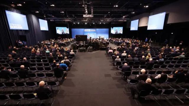 Wide shot inside the conference room, showing a speckling of people sitting on chairs watching on, but there are also a lot of chairs empty