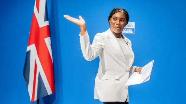 Badenoch waves as she walks across a stage, wearing a white suit jacket and white top underneath.