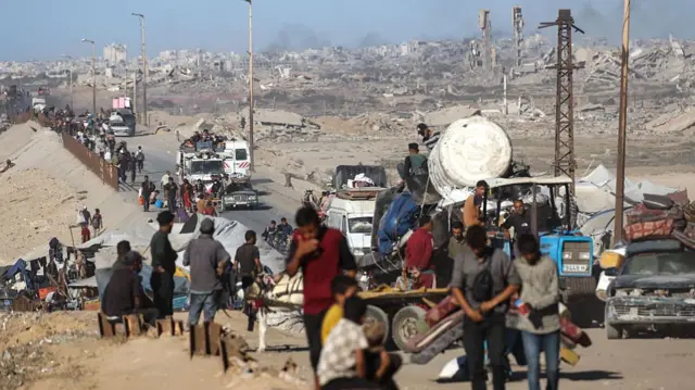 crowds of Palestinians walk along road as smoke rises from distant buildings
