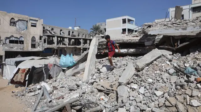 a child stands on top of a large pile of building rubble surrounded by destroyed buildings