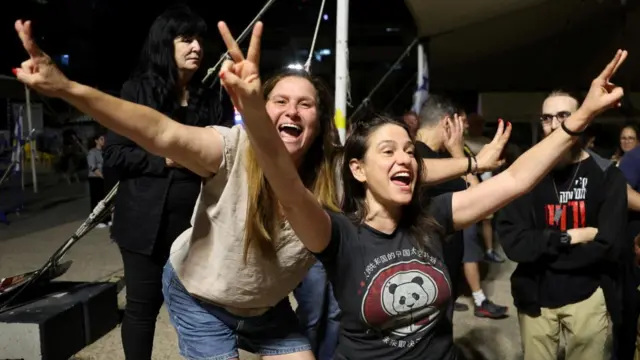 Two women with wide smiles and arms in the air celebrating and making the peace sign, with others standing nearby