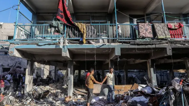 two boys grab hands as they step over rubble in front of damaged building