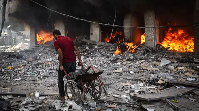 man drags wheelchair behind him through rubble as flames and smoke rise from building