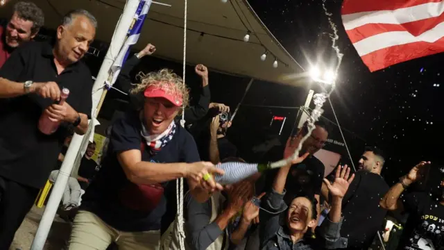 A woman, smiling and holding a bottle with champagne shooting out of it with others celebrating behind her and a partial US flag