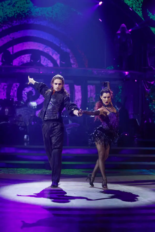 George and Alexis hold hands as they walk towards the audience, and they're other hands are splayed out to the side. They're dressed in purple outfits, and don serious expressions