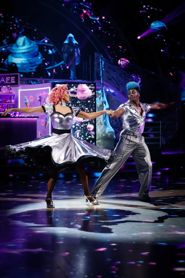 Harry and Karen wear shiny, silver ensembles and have blue and pink dyed hair, respectively. They splay their arms out in a flourish, as they hold hands and point their toes to the side