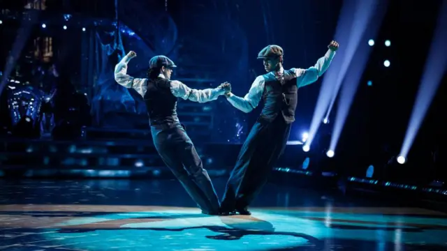 Peaky Blinders dressed dancers hold each others' hands as they pull their bodies away from one another. They're both wearing flat caps