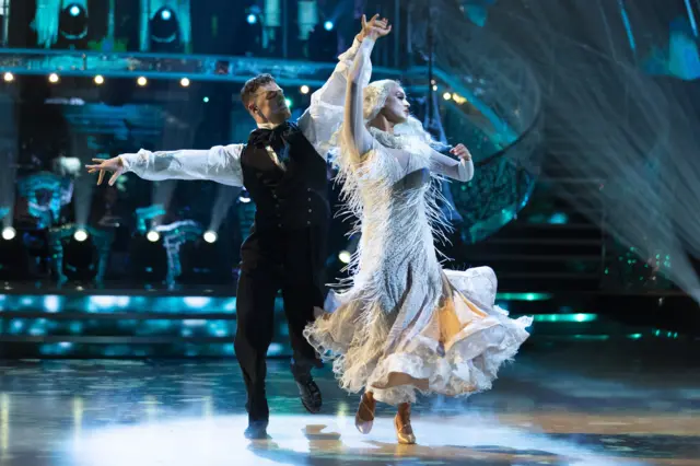 Vicky and Kai waltz across the floor, with blue lights shining down above them. Vicky wears a flowy white dress, while Kai wears a black vest with a white dress shirt underneath that has puffy sleeves.
