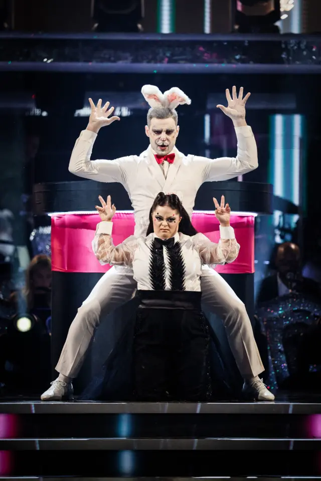 Ellie, dressed as a magician, poses below her partner Vito, who is dressed as a rabbit. They put their hands up like claws and wear semi-serious expressions