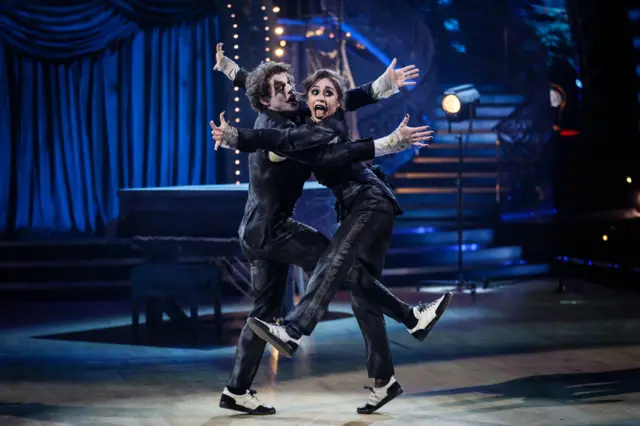 Lewis Cope and partner Katya Jones wear black pants and jackets and wear joker-esque face paint. They're bodies are intertwined, and they wear animated expressions as their arms and legs collide