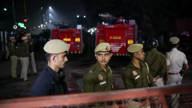 Several men in uniforms stand by a fence in front of two fire engines