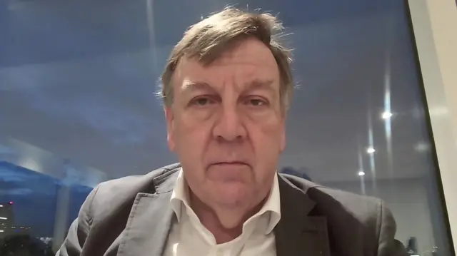 Sir John Whittingdale sits in front of a window with a view of the dark early morning sky wearing a grey blazer and open collar shirt