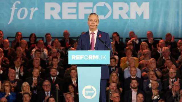 Nigel Farage stands on a pedestal marked with the Reform UK logo. He's surrounded by an audience, applauding.