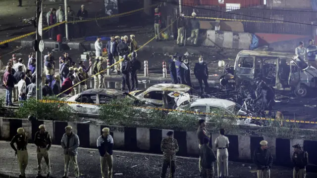 general A row of emergency service personnel stand in front of several burnt out vehicles. There are at least three cars and a minibus among the wreckage. To the left of them is police tape with a crowd of people standing