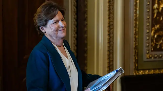 Sen. Jeanne Shaheen, smiling and wearing a blue blazer and white blouse, holds a folder as she walks