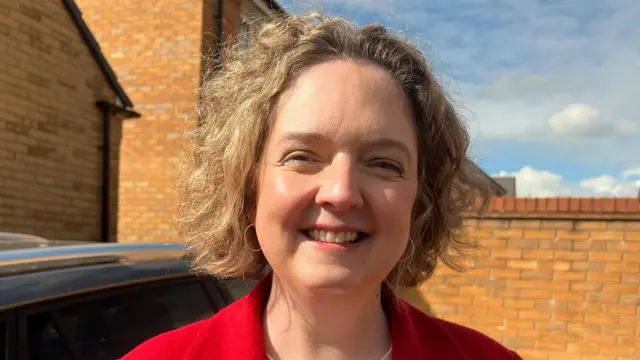 Anna Sabine is smiling outside a new build estate. She is wearing a red blazer.