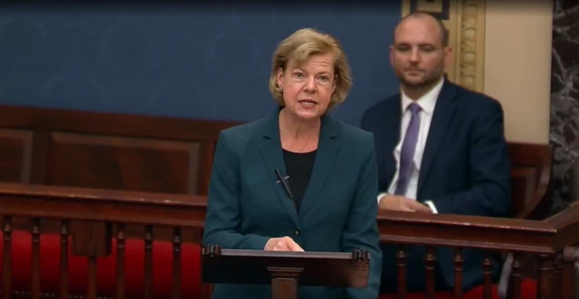 Tammy Baldwin, wearing a dark green suit over a black shirt, speaks on the senate floor