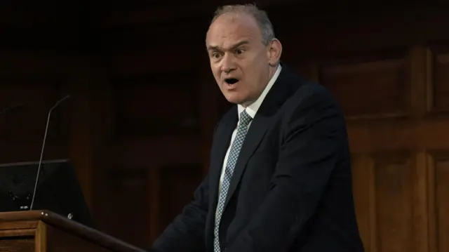 Ed Davey stood at a pedestal performing a speech. He looks passionate and is wearing a dark suit with a light blue, patterned tie.