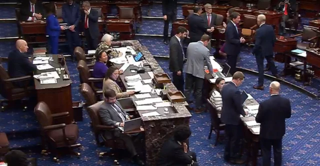 People standing around and sitting at desks with a blue carpet and wooden furniture