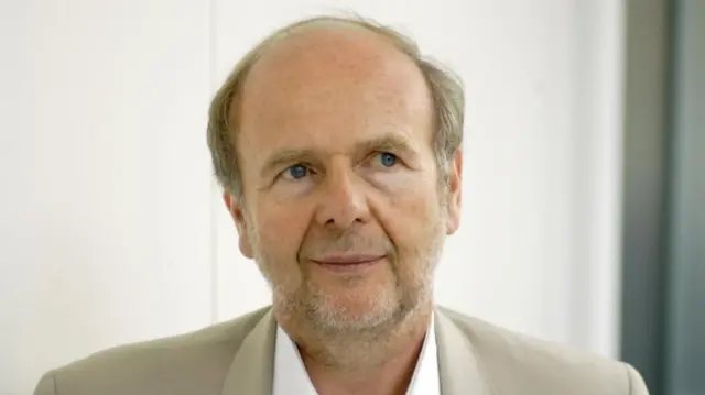 a man in a cream jacket stands in front of a white wall
