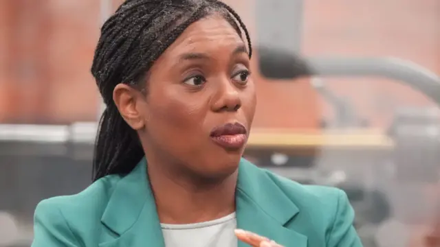 Kemi Badenoch in a studio speaking to a interviewer off camera. She is wearing a blue/green blazer on top of a white top.