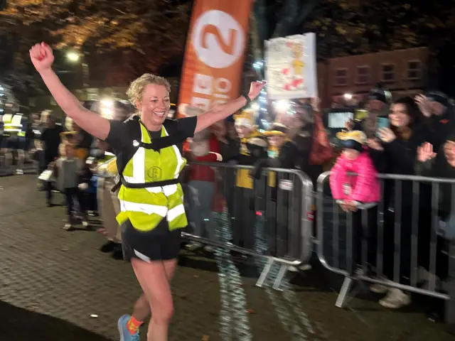 Sara Cox running across the finish line for Day 2, she's smiling with her arms in the air