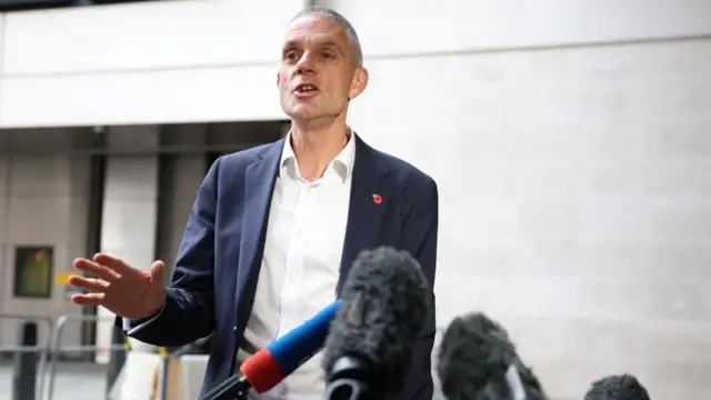 Tim Davie, in a suit and open collar shirt, addresses microphones outside Broadcasting House